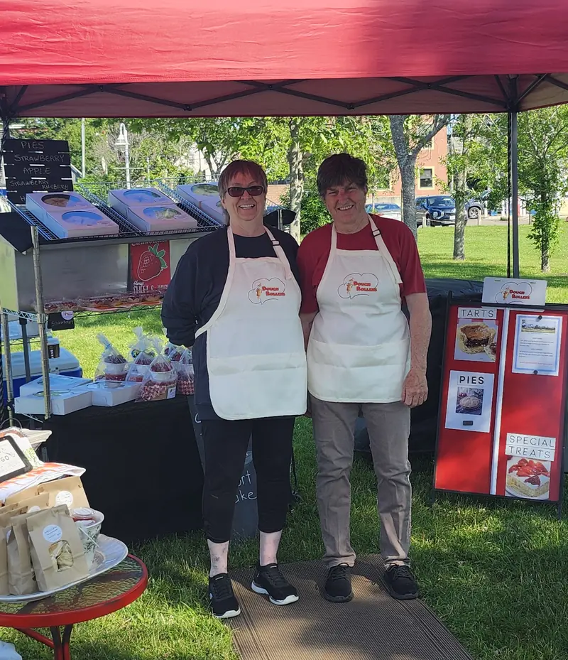 Cathy and Jan in Dough Rollers aprons under their red market tent, with pies and treats on the tables
